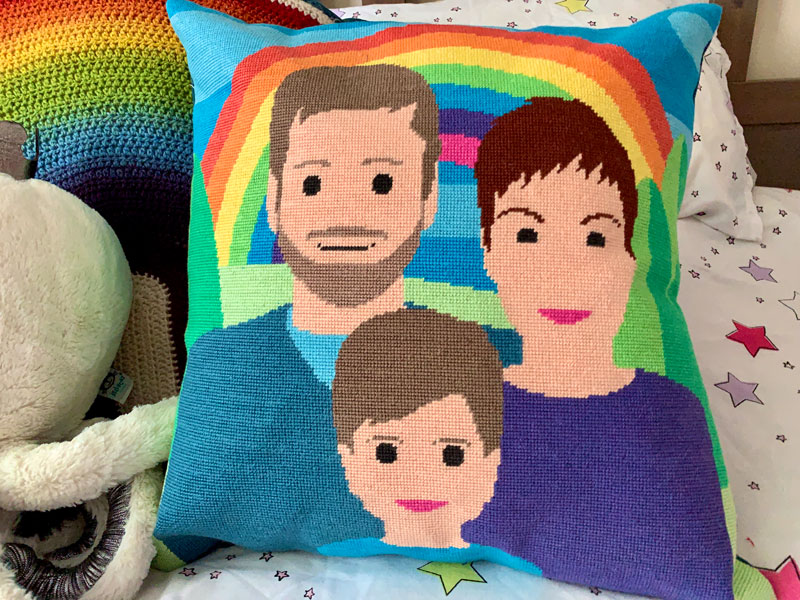 a handmade tapestry cushion on a bed. The image on the cushion is a stylised family portrait in front of a rainbow.