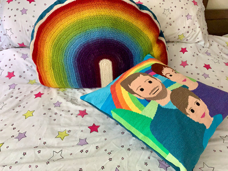 a handmade tapestry cushion on a bed. The image on the cushion is a stylised family portrait in front of a rainbow.