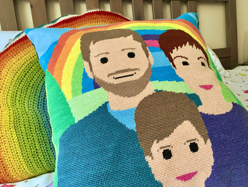 a handmade tapestry cushion on a bed. The image on the cushion is a stylised family portrait in front of a rainbow. 