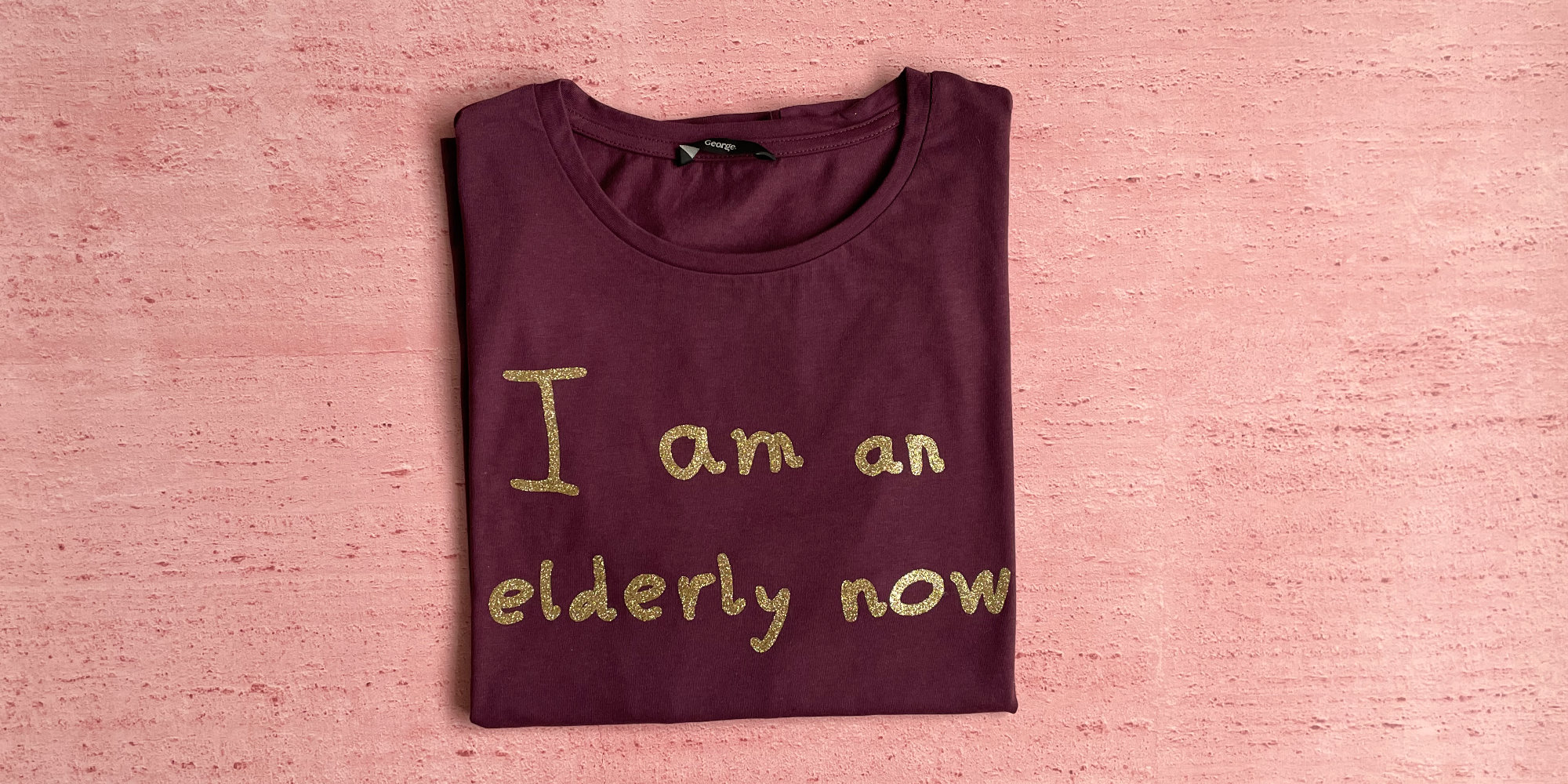 A folded maroon t-shirt with a child's handwriting on it, in gold, reading 'I am elderly now'.