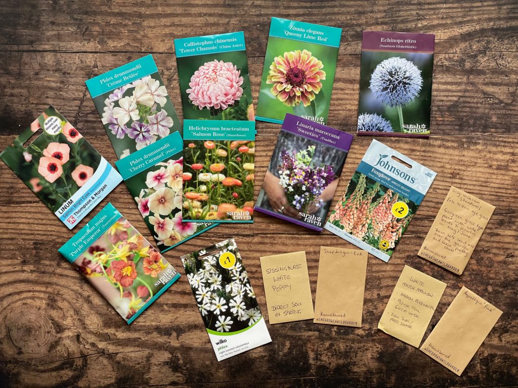 Am old wooden table top, top down, with packets of seeds: Line, Phlox, tower chamois, zinnia, chinos retro, nasturtiums, strawflowers, foxglove, poppies. Other seed packets are obscured/too small to read.