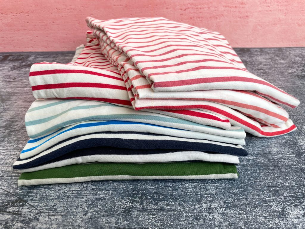 Pile of stripy tops in multiple colours on a slat table with a pink wall in the background,. The tops are folded and the pile is slightly lopsided.