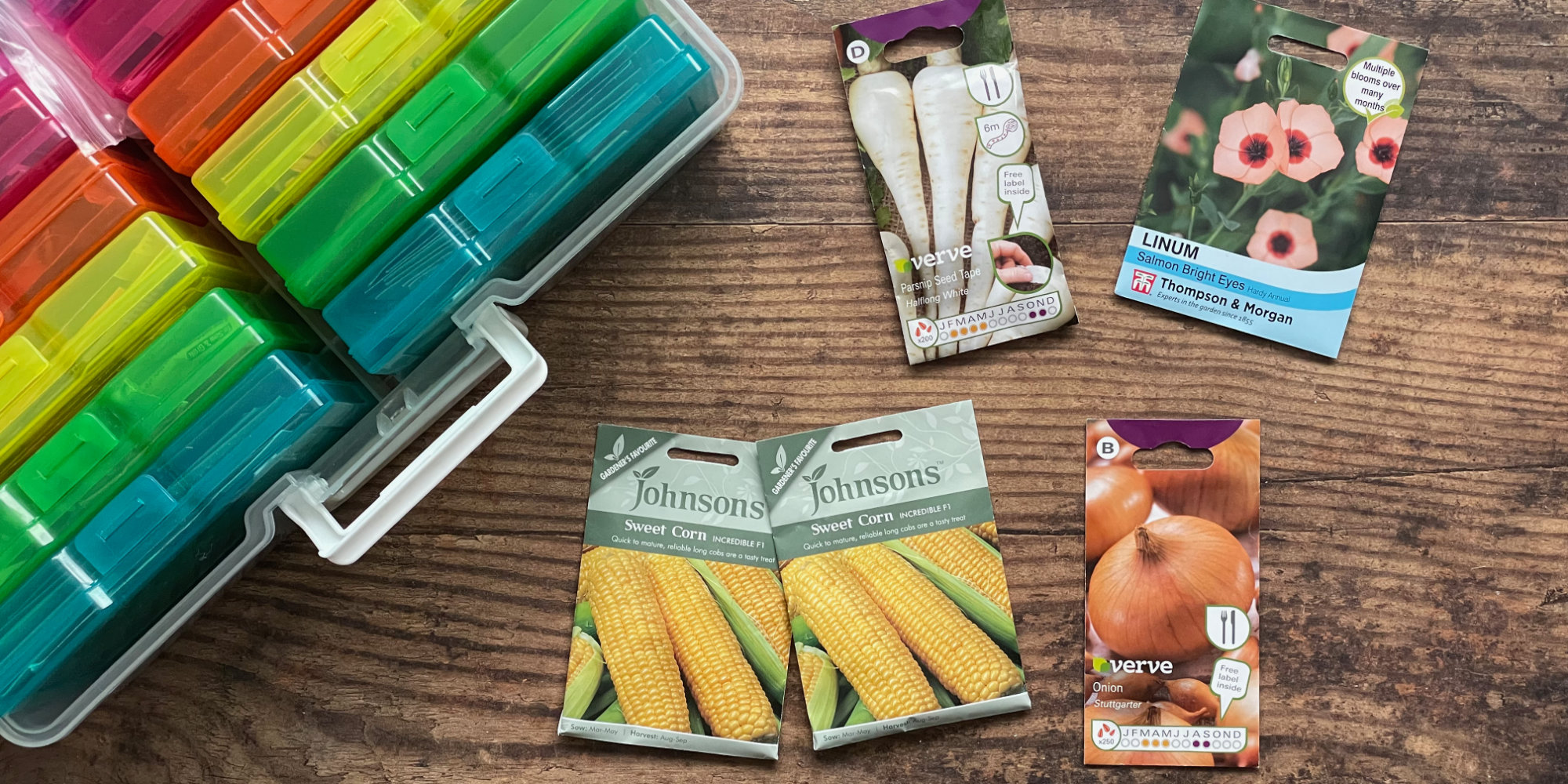 an open clear plastic case, containing 10 visible, smaller, plastic inner cases in different colours is open in the top left corner of the photo. Multicoloured packets of seed (sweetcorn, onions, parsnips and flowers) are next to the case, and it all sits on a scarred wooden table.