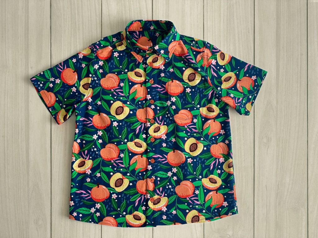 Front view of a shirt made from a bold printed fabric of peaches and leaves on a blue background. The shirt is buttoned up and has short sleeves.
