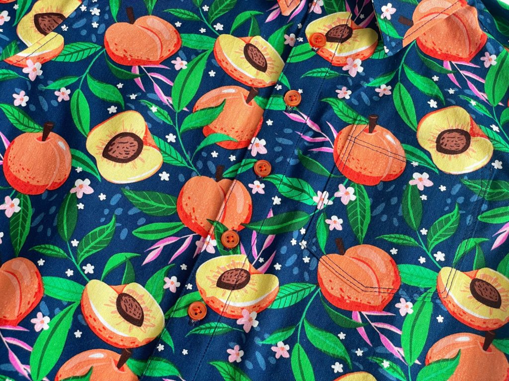Close up of a shirt showing the button bands and pocket. The shirt is make from a bright cotton print showing peaches and leaves on a blue background. 